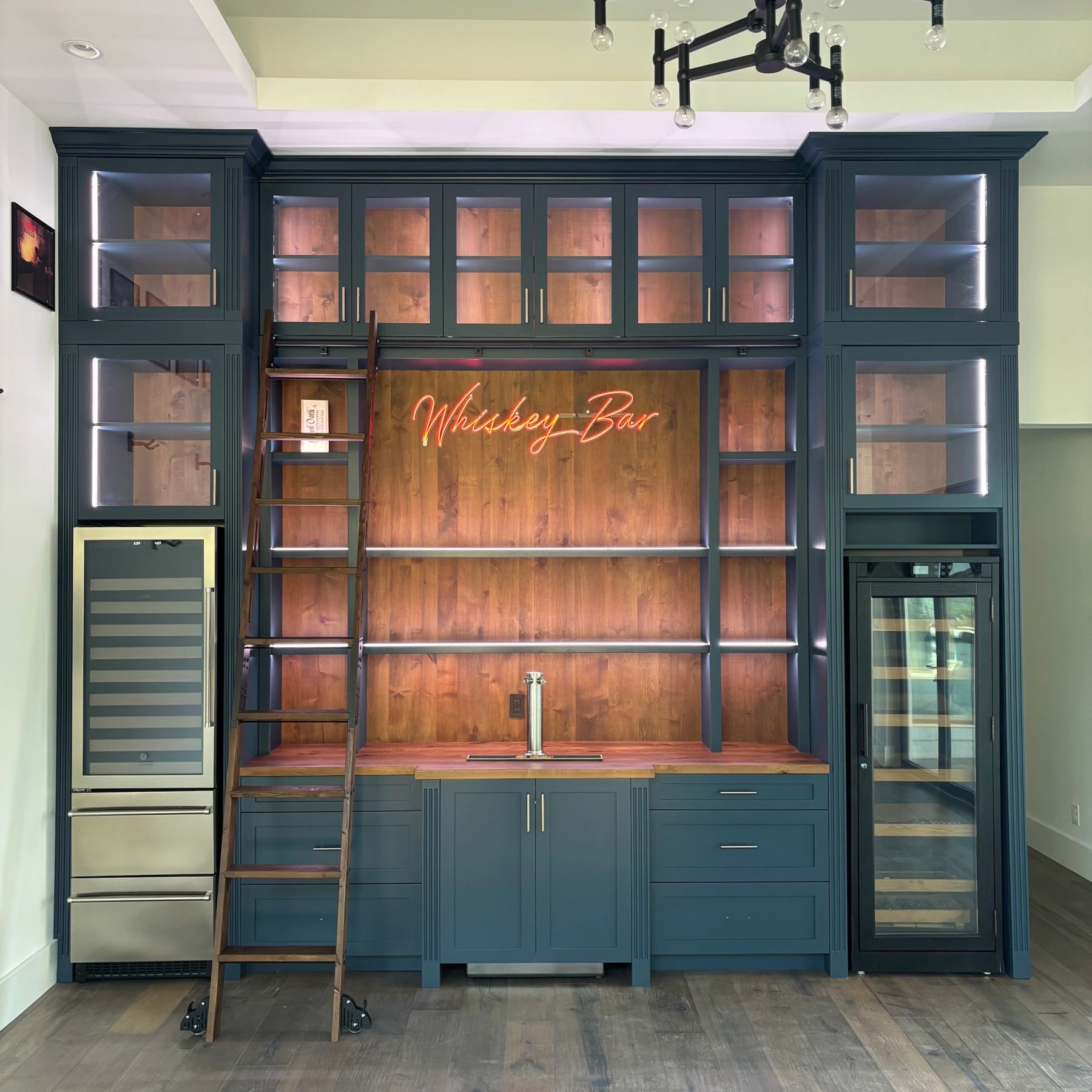 Built-in whiskey bar with deep blue cabinetry, rolling ladder, and lighted glass display cases by Westcreek