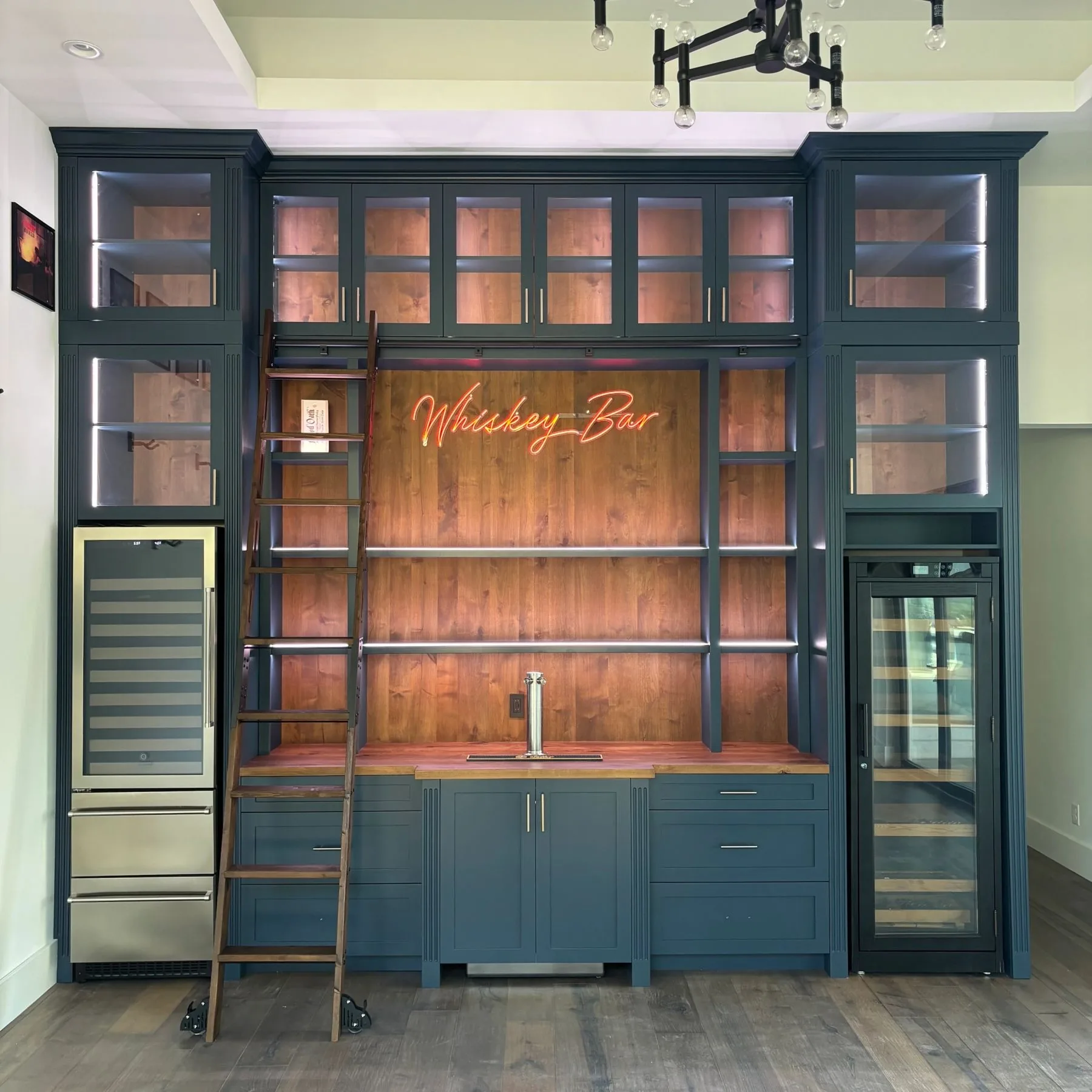 Built-in whiskey bar with deep blue cabinetry, rolling ladder, and lighted glass display cases by Westcreek